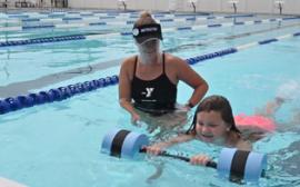 swim instructor standing in the pool next to child swimmer, holding floating barbell