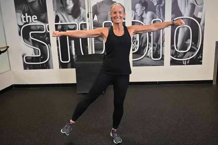 Exercise instructor smiles while posing with arms in a T and raised leg.