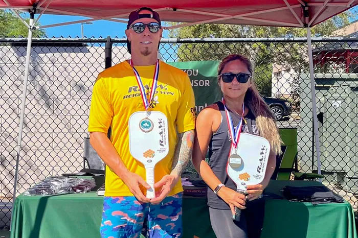 Tall man next to woman, both wearing sunglasses posing in the sunny outdoors with their pickleball tournament medals and paddles.