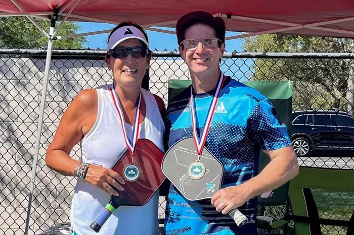 Woman and man stand in the sunny outdoors, under a tent. They are wearing medals and pickleball paddles from the 2023 St. Paddles Day Pickleball Tournament.