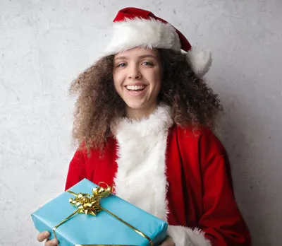 Tween femail in Santa suit holding blue gift box with gold bow. Gray backdrop, stronger lighting on left side.