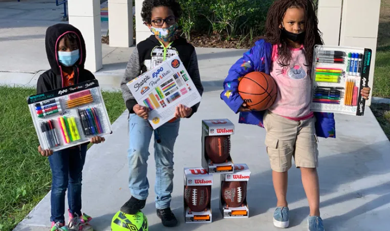 three children holding school supplies and sports balls outside