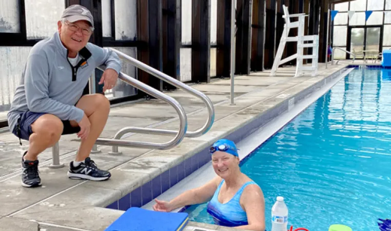 senior woman in pool smiling, with male coach squatting for photo