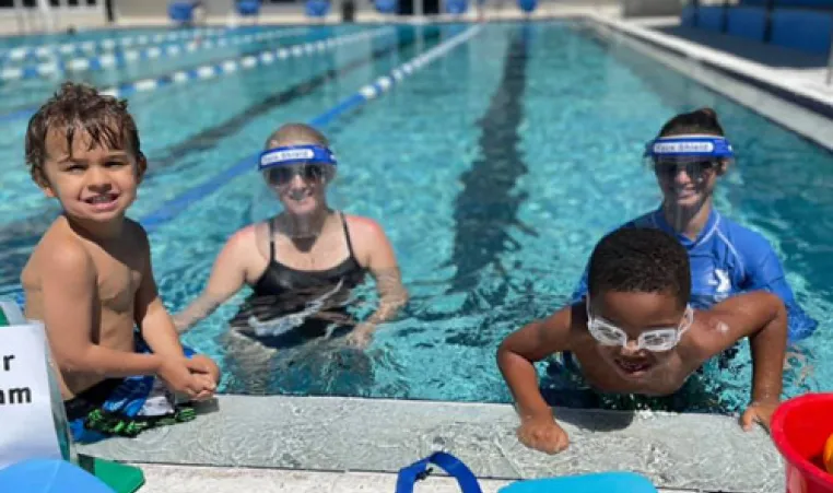 Two swim instructors wearing face shields standing in pool with two young kids sitting on edge of pool during swim lessons