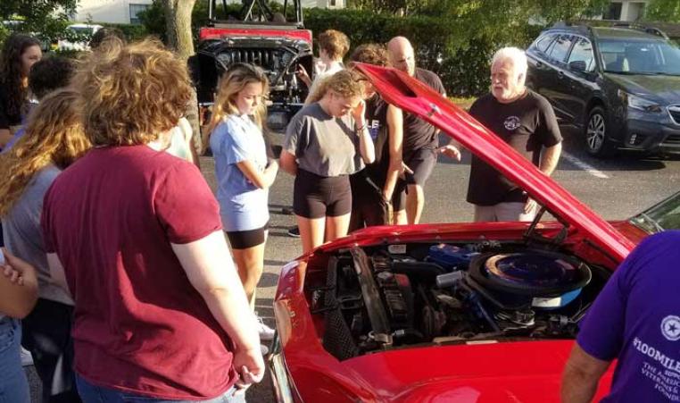 A group of teens from Teen Leaders Club at John Geigle gather around a red car with the hood lifted. Car aficionado and YMCA namesake John Geigle teaches the group about caring for cars. 