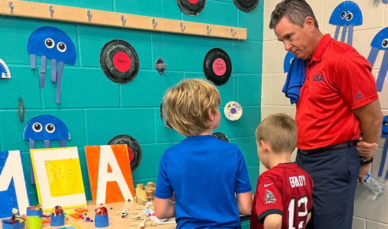 Man standing at craft display table with two children as they show off their work.