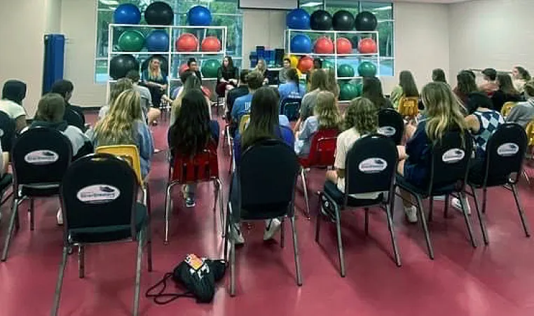 Teens in a large room with red floors, sitting in chairs listening to adult speaker during a meeting.