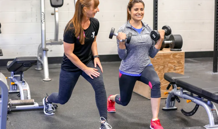 Female personal trainer demonstrates a forward lunge to female client, as client is in the stance. Client is holding dumbbells in bicep curl position.