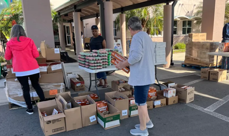 Four volunteers at James P. Gills YMCA organize food donations outside in the shade. Many brown boxes of food rest on pallets and the ground. Foreground includes a man facing away from the camera, holding cereal boxes.