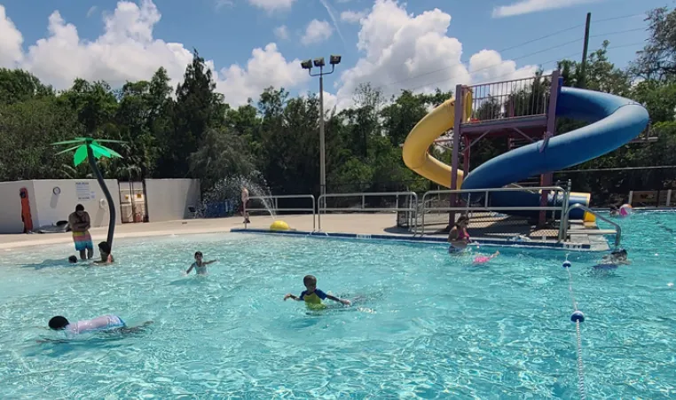 Kids playing in outdoor zero-entry pool with waterslides in the background. Sunny day with puffy white clouds and tall evergreen trees in background scenery.