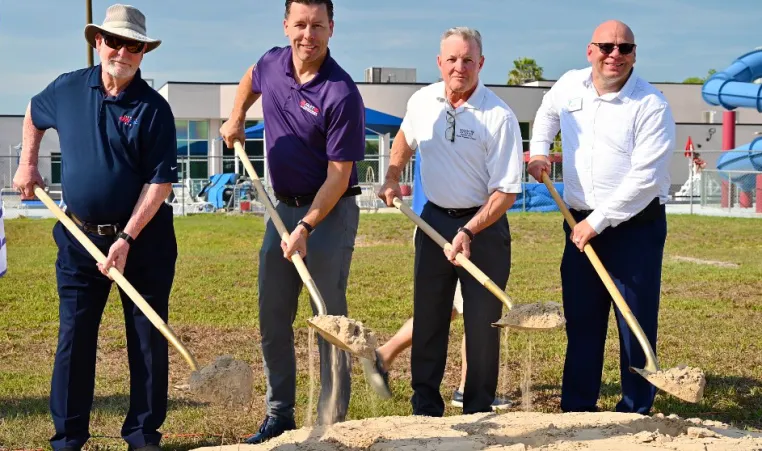 Outdoor image of four men posing for a groundbreaking photo. The subjects are holding shovels with dirt. A swimming pool with slides is in the background.