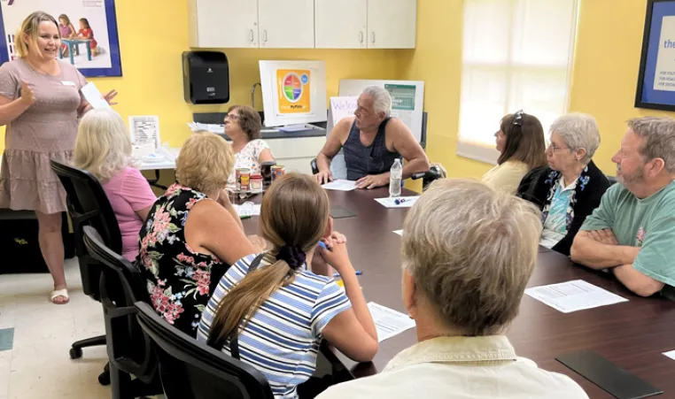 A group of nine adults sitting at a table, listening to a woman standing, teaching the class. Background: classroom walls are yellow.