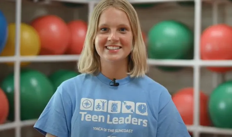 A teen girl with short blonde hair and a light blue Teen Leaders YMCA shirt smile at the camera. The background is out of focus, and shows white shelves holding green and red exercise balls.