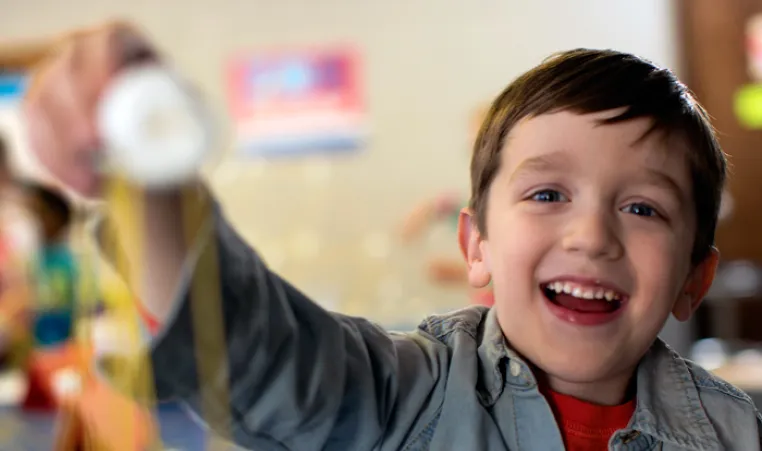 A young boy smiles as he reaches his arm out to show off the craft he made. The child's face is in focus and the craft is in the foreground, out of focus.