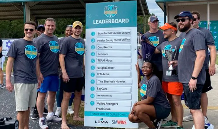 10 men and women pose by Corporate Cup Leader Board as winners for the day 