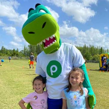 Two children at 30th Healthy Kids Day® pose for a photo with Publix mascot, Plato the Publixaurus. Background: kids playing sports and inflatable obstacle course.