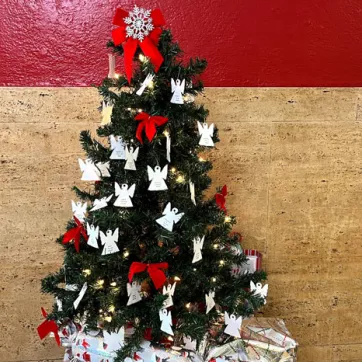 A small Christmas tree with paper angel ornaments and red bows in front of red plus tan stone wall.