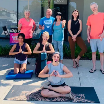 YMCA staff and members smiling outside new facility. Three people are on the ground in yoga pose with prayer hands and five others stand behind them.