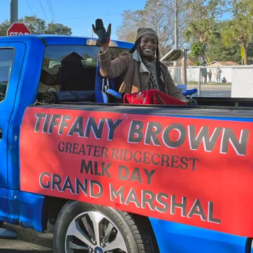 Ridgecrest leader waves from the bed of a blue truck for Greater Ridgecrest MLK Day parade. Sign is on side of truck bed, black block letters in all caps: Tiffany Brown Greater Ridgecrest MLK Grand Marshal on red background.