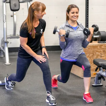 Female personal trainer demonstrates a forward lunge to female client, as client is in the stance. Client is holding dumbbells in bicep curl position.