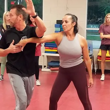 Man and woman instructors demonstrate self defense move to teens in background.