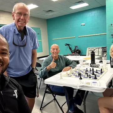 4 members of Chess Club smiling for photo. Two are sitting beside the chess table.